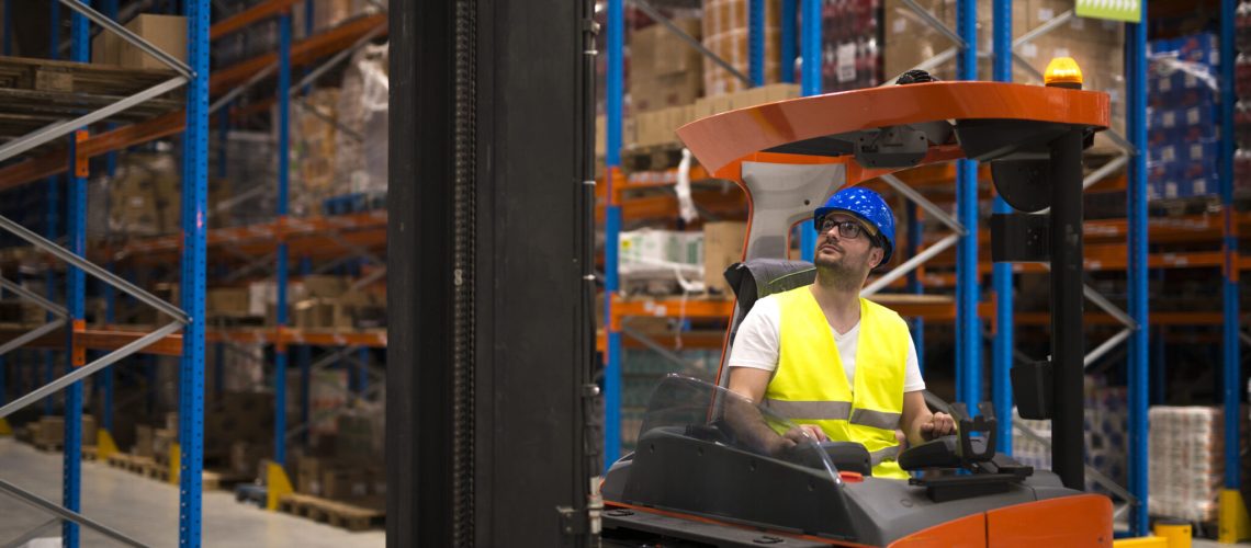 Forklift driver relocating and lifting goods in large warehouse center.